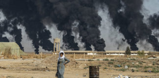A woman stands along the side of a road on the outskirts of the town of Tal Tamr near the Syrian Kurdish town of Ras al-Ain along the border with Turkey in the northeastern Hassakeh province on October 16, 2019, with the smoke plumes of tire fires billowing in the background to decrease visibility for Turkish warplanes that are part of operation "Peace Spring"