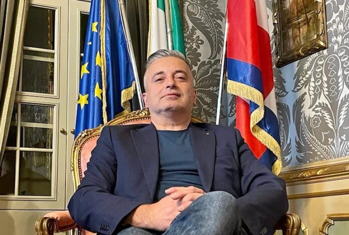 Graglia Franco seduto con le gambe incrociate davanti alla bandiera dell'Europa e della Regione Piemonte