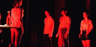 illuminati da un faro rosso quattro attori in piedi intorno a una ragazza non illuminata prostrata a terra